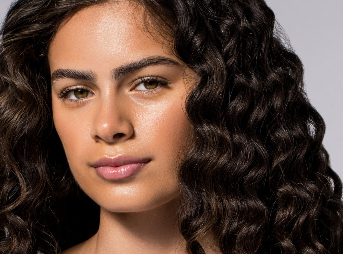 A close-up portrait of a woman with long, dark brown, tightly curled hair against a light gray background.